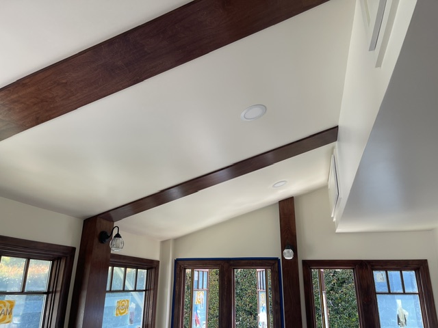 Wooden ceiling beams and recessed lights in a room with large windows and white walls; sunlight is coming through the windows.