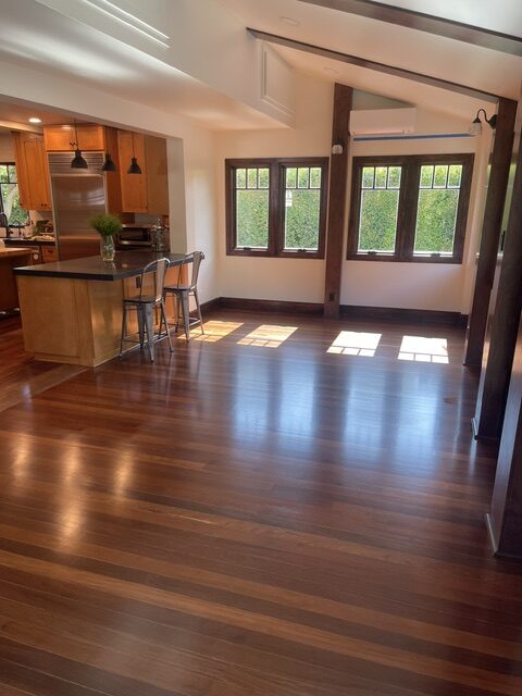 Sunlit room with dark wood floors, large windows, and an open kitchen area featuring an island with two stools and pendant lights.