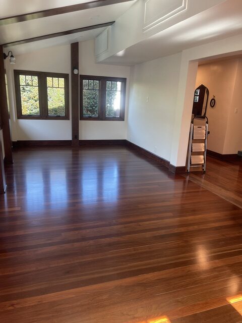 An empty room with polished wooden floors, white walls, large windows with wooden frames, and a small stepladder near an arched doorway.