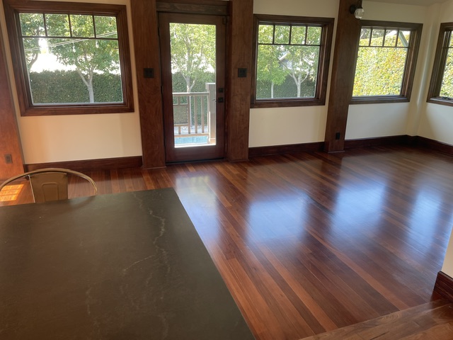 Sunlit room with polished wooden floors, large windows, a door leading outside, and a black tabletop in the foreground.