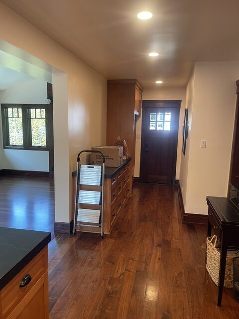 A hallway with wood flooring features a small set of steps, a cabinet with boxes, and a dark wooden door at the end; an adjacent room with windows is visible to the left.