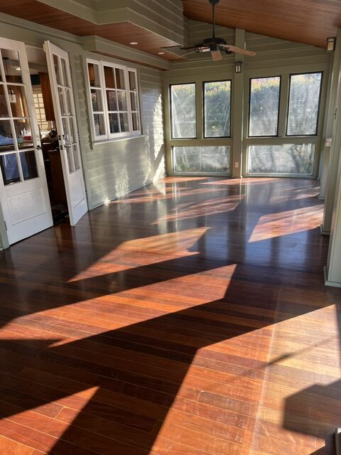 Sunlight streams through multiple large windows onto polished hardwood floors in an empty, enclosed sunroom with green walls and white trim.