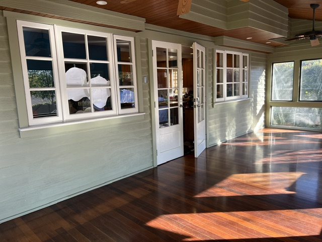 A sunlit room with green walls, white-trimmed windows, double glass doors, and dark wood flooring. Sunlight casts shadows across the floor.