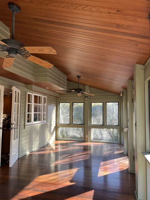 A sunroom with green walls, wood ceiling, ceiling fans, large windows, and polished wood floors. Sunlight streams in through the windows, creating bright patches on the floor.
