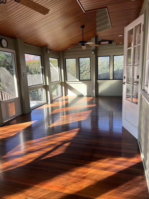 Sunlight streams through large windows onto the polished wooden floor of an empty sunroom with a wood-paneled ceiling and ceiling fans.