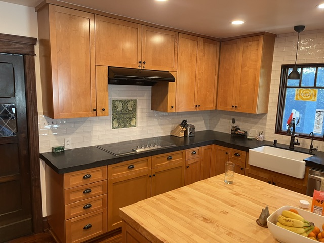 A kitchen with wood cabinets, black countertops, a farmhouse sink, an electric cooktop, and an island with a wooden top. Fruit, utensils, and a glass are on the island.