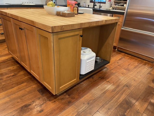 A wooden kitchen island with a lower open shelf holding storage containers, situated on a polished wood floor next to stainless steel appliances.