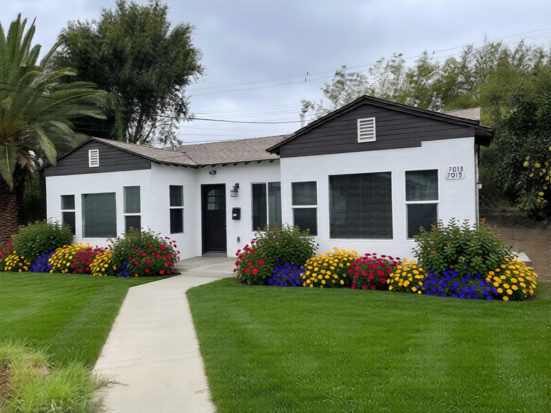 Awhite ADU with dark trim, surrounded by green lawn and colorful flower beds, under a cloudy sky.